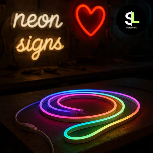 Strip LED neon RGB accesa appoggiata su un bancone di un laboratorio, con insegne neon luminose sullo sfondo e logo StripLed.it