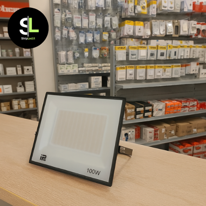 Faro LED 100W esposto su bancone di negozio con scaffali di prodotti elettrici sullo sfondo e logo StripLed.it”