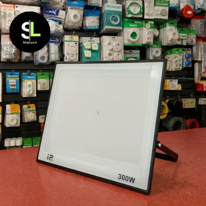 Faro LED 300W esposto su bancone di negozio con prodotti elettrici sullo sfondo e logo StripLed.it