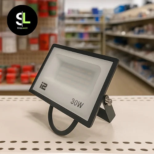 Faretto LED 30W esposto su bancone di negozio con scaffali di prodotti elettrici sullo sfondo e logo StripLed.it